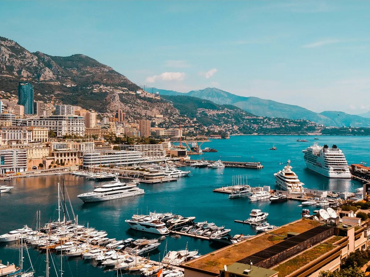 A picturesque coastal cityscape with yachts moored in a marina, framed by mountains under a clear blue sky.