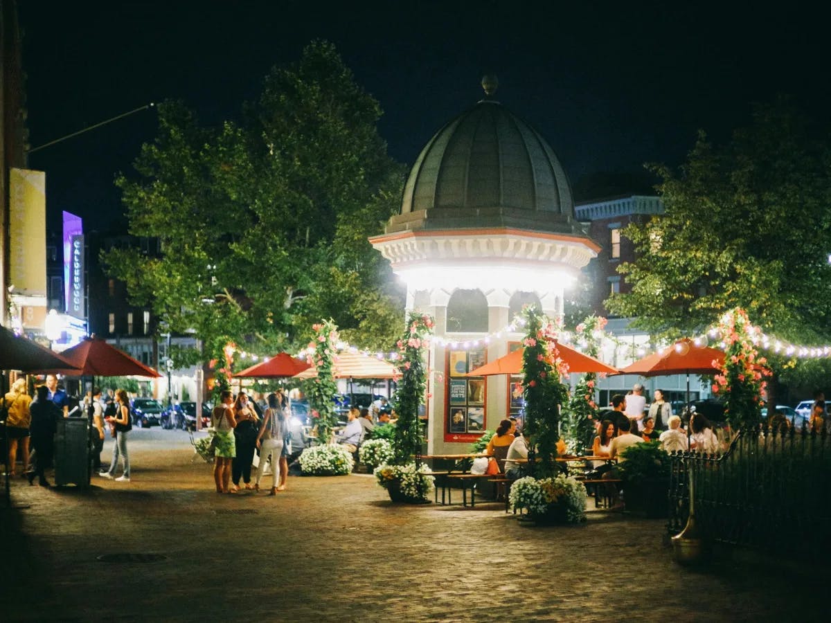 A lively outdoor evening scene with people gathered around a central kiosk under ambient lighting.