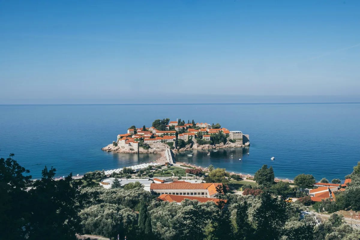 Sveti Stefan, a picturesque islet and luxury resort off the coast of Montenegro, with a rocky island with red-roofed buildings on it off of the coastline.