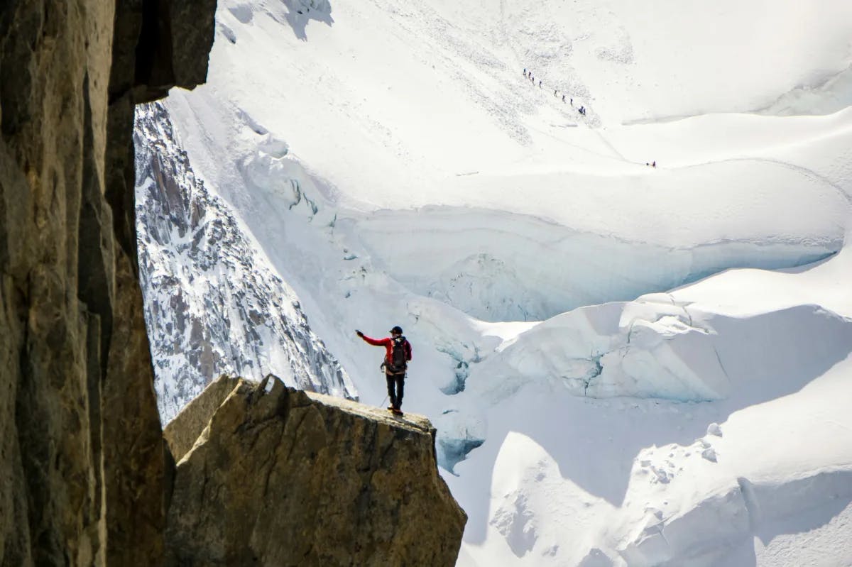 A person standing at the edge of a slanted cliff while wearing outdoors gear and looking out onto a large snowy mountain.