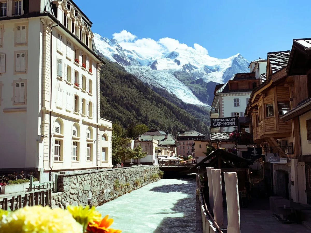 A view of a canal surrounded by stone ledges, luxurious buildings and lodges, and a view of the green and snowy mountains in the distance.