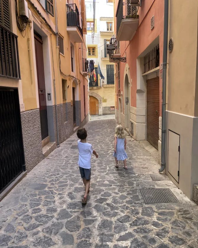 Two children walking down a quaint, narrow street in between red and orange colored buildings