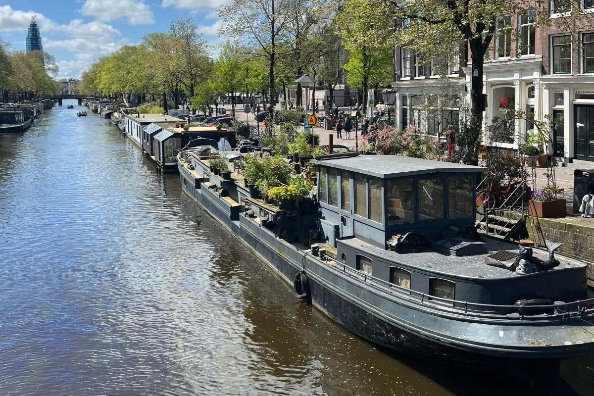 A row of riverboats docked along one of Amsterdam's canals, with shops, buildings and people walking down the banks of the canal.