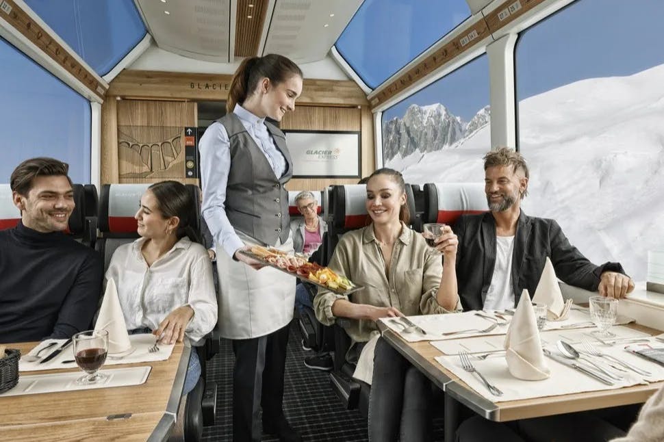 An image of people dining inside of a train car with views of snowy hills in the surrounding area. 