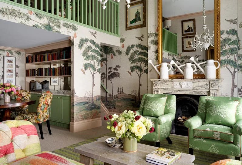 a sunlit living room with green upholstered chairs amid wallpaper displaying a verdant landscaoe