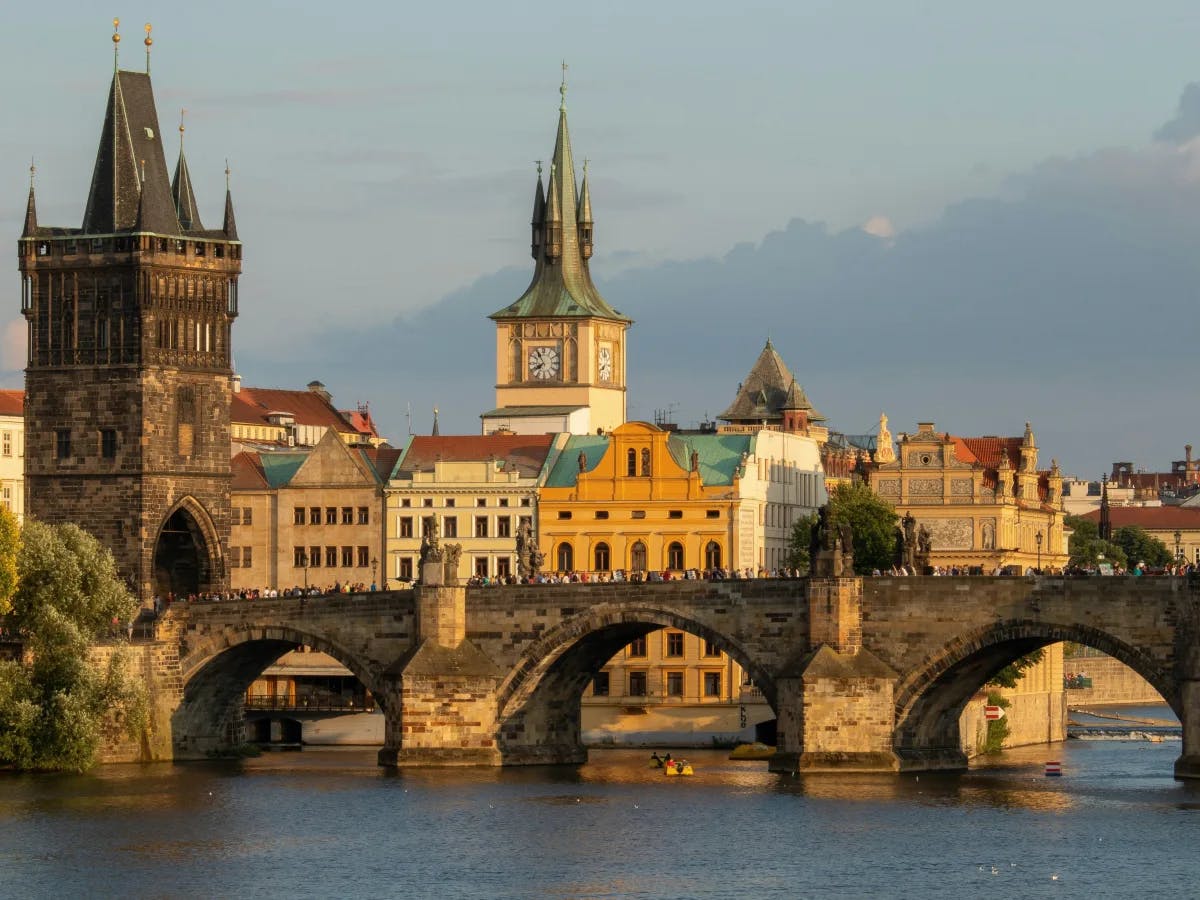 The image presents a historic stone bridge with towers, crossing a river against a backdrop of charming old buildings and a clear sky.