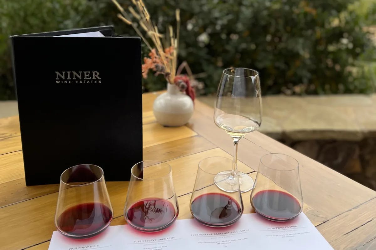 A flight of red wines and one white on a wooden table next to a Niner menu on a patio.