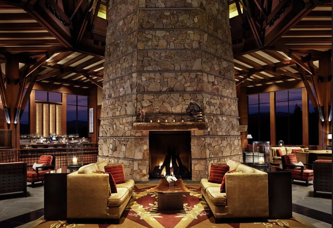 An interior view of a hotel lounge with a beautiful stone fireplace, wood paneled ceiling, luxurious furniture and lots of accent lighting. 