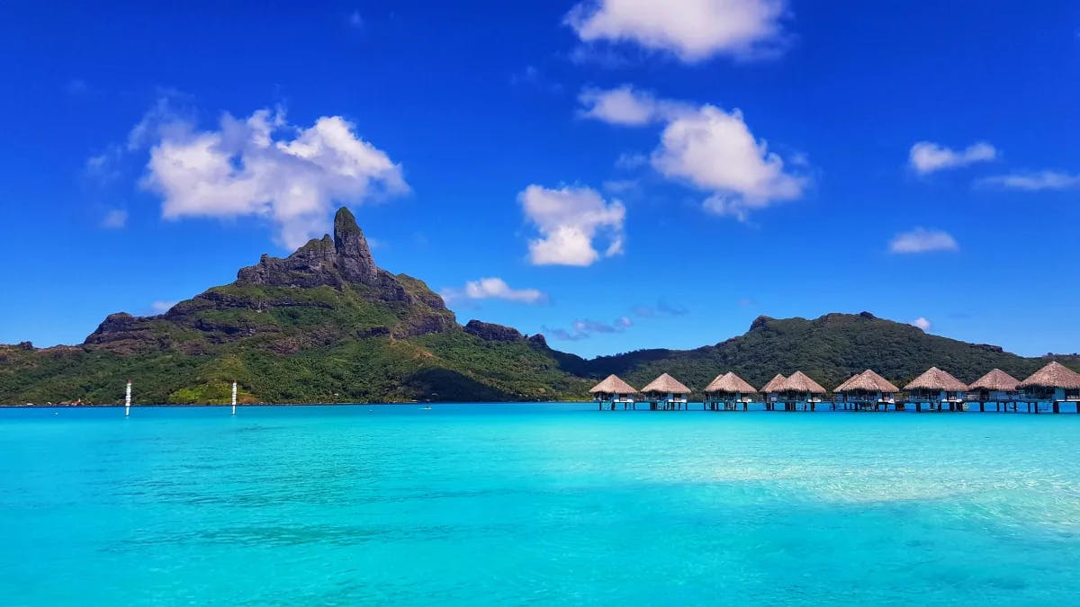 A view of Bora Bora with huts over the water and a mountain.