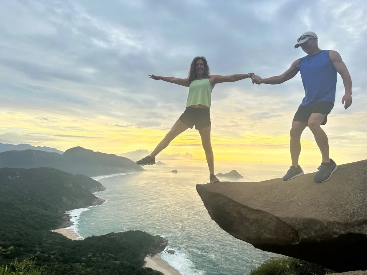 A man and woman posing on the edge of a cliff overlooking the ocean at sunset. 