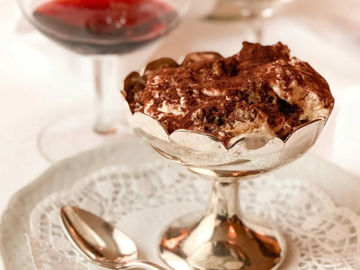 An image of tiramisu in a silver cup placed atop of a doily resting on a white plate with a silver spoon on the left side and a glass of red wine in the background. 