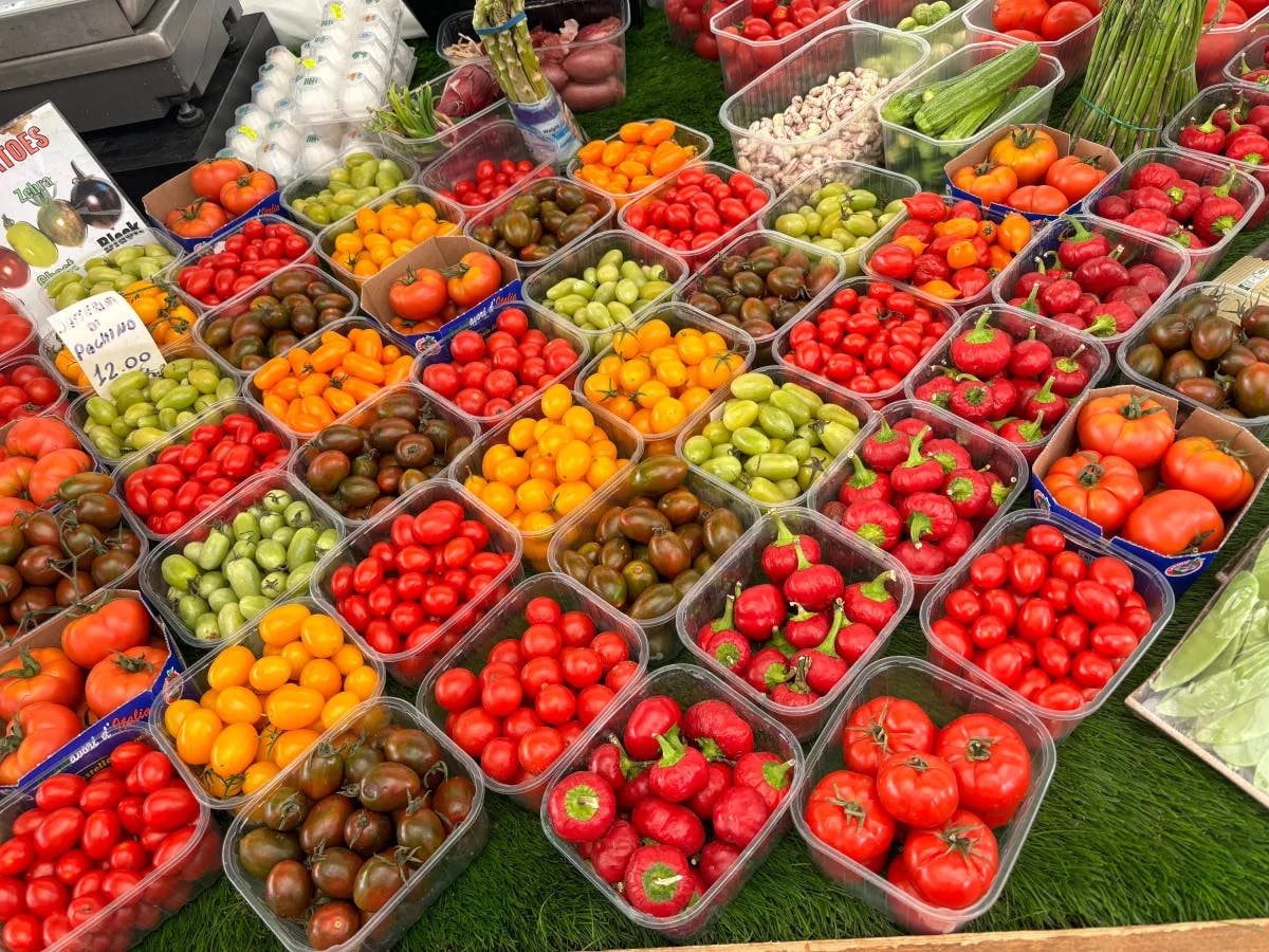 The image features a vibrant display of fresh vegetables in clear plastic containers at a market.