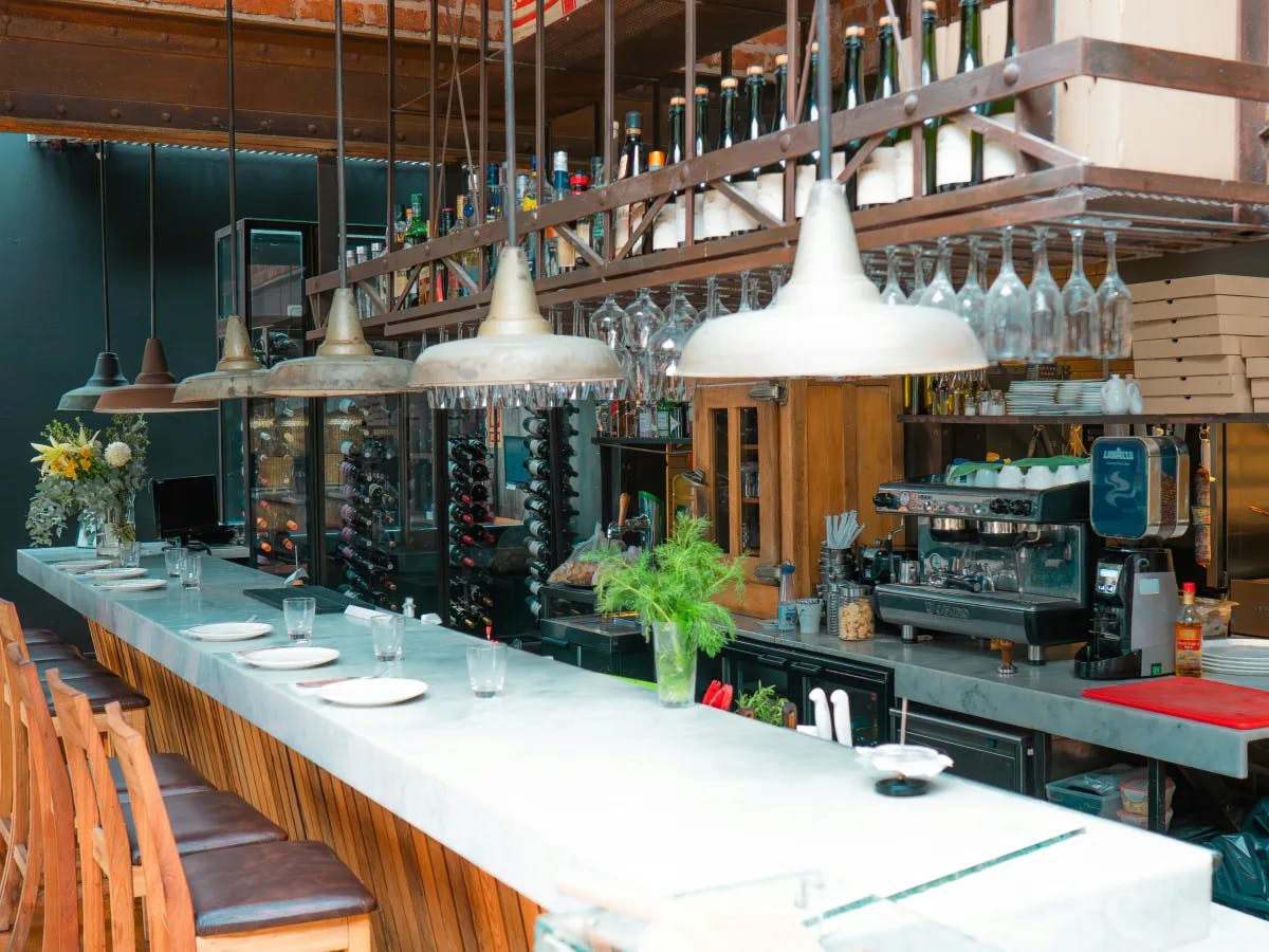 The image shows an interior view of a bar or restaurant with a long counter, bar stools, hanging lights, and shelves stocked with bottles and glasses.