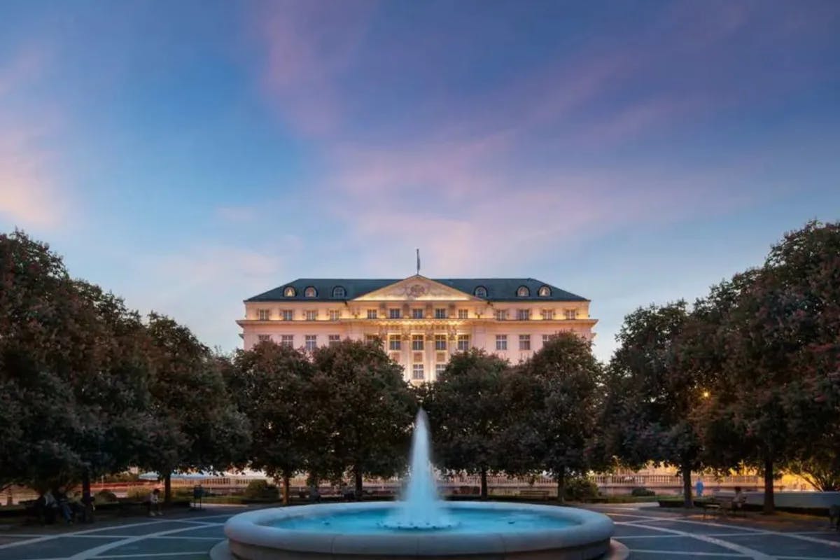 fountain with surrounding trees at dusk with regal hotel facade in the background