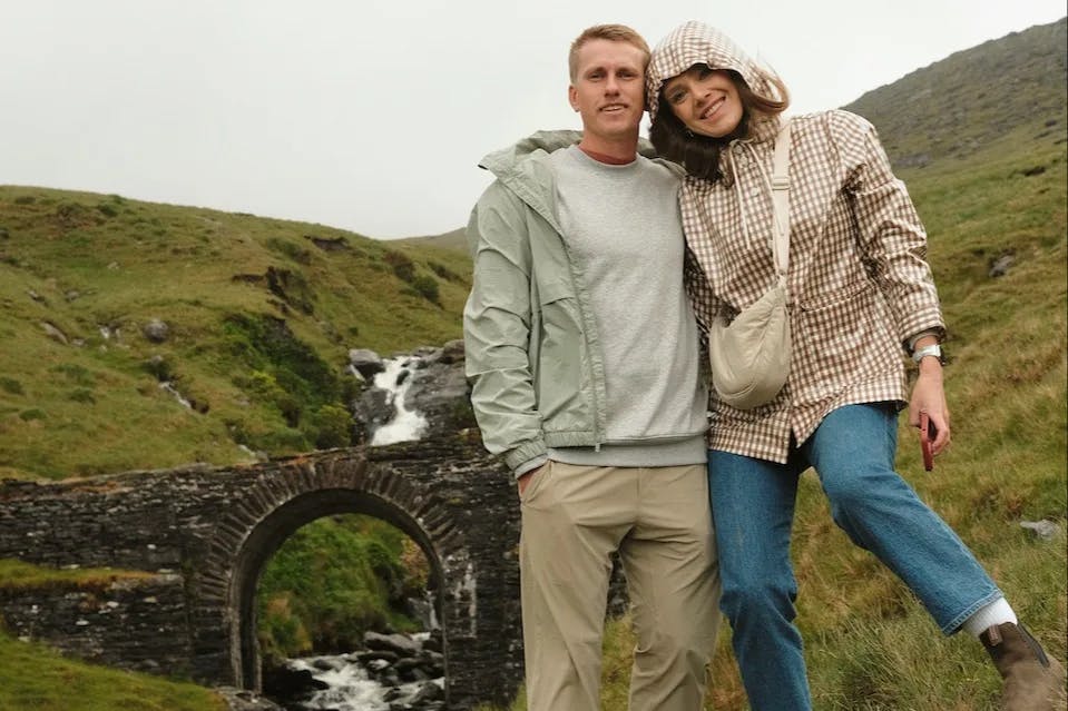 A man and woman pose together in front of a water fall and bridge between two green rolling hills at Healy Pass waterfalls