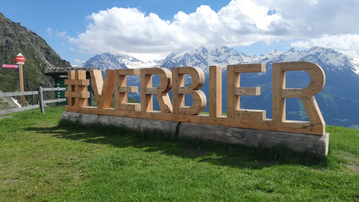 A signage with letters displaying Verbier with snow-capped peaks in the distance on a sunny day dotted with rolling clouds.