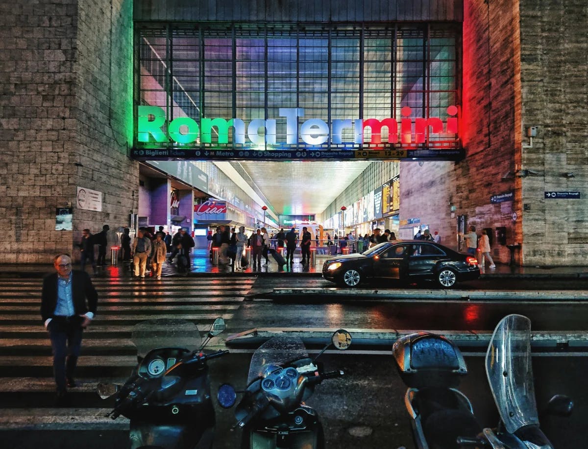 A neon sign reading, "Roma Termini" sits atop a brick building lit up by the bustling crowds and nightlife. 