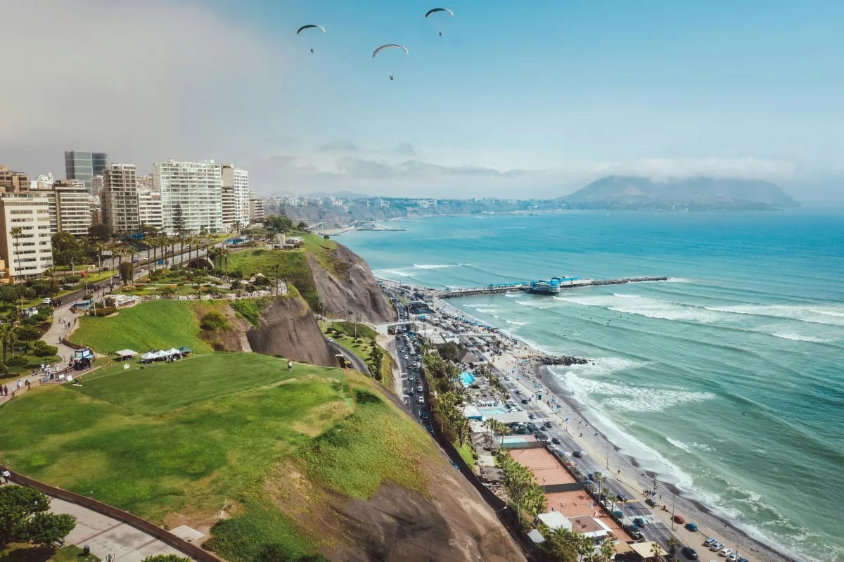 An aerial view of Lima, Peru with the city skyline, coastline, azure water, people parasailing and mountains in the background. 