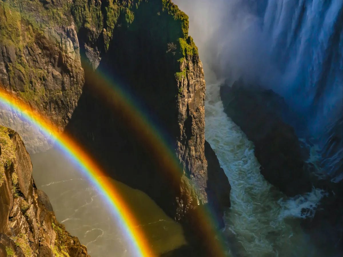 An overhead view of a rapid river, waterfall and rocky, green cliffs with a beautiful double rainbow shining from above. 