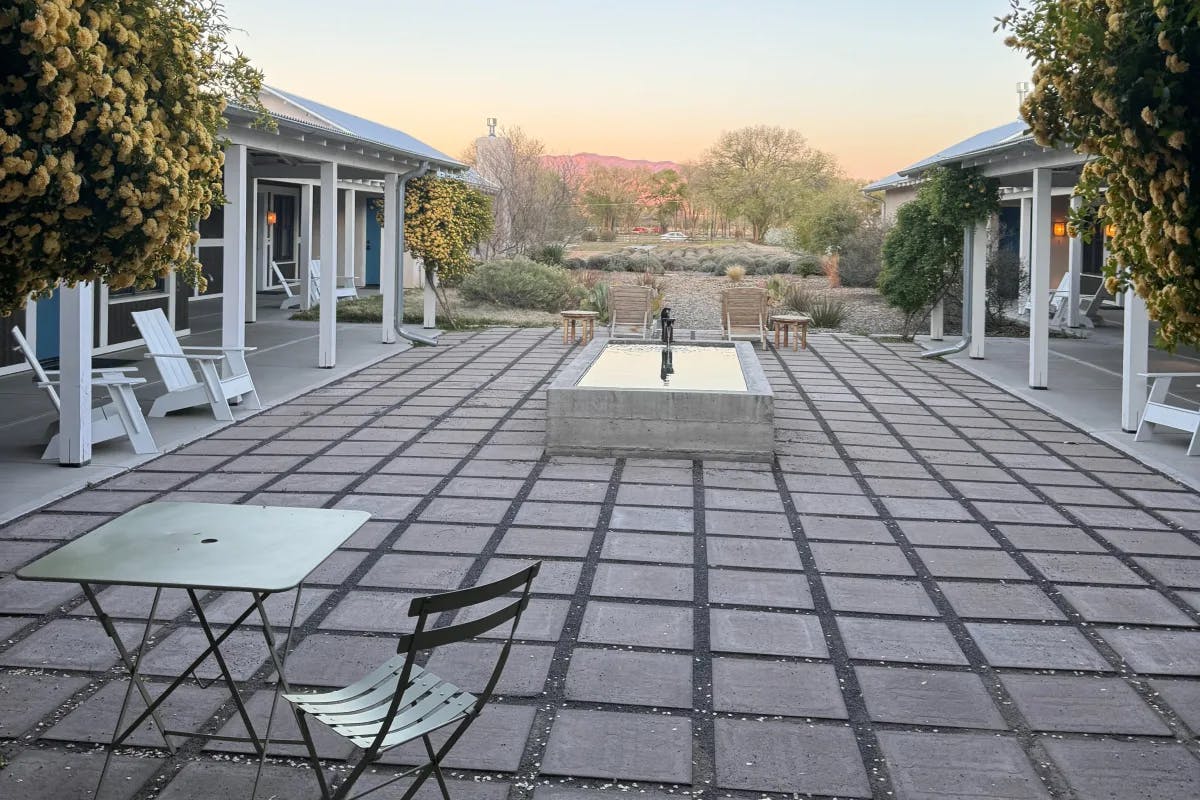 This image depicts a stone fountain in the middle of a garden with bistro tables, white pergolas, beuatiful landscaping, floral trees and white lounge chairs in the surrounding view. 