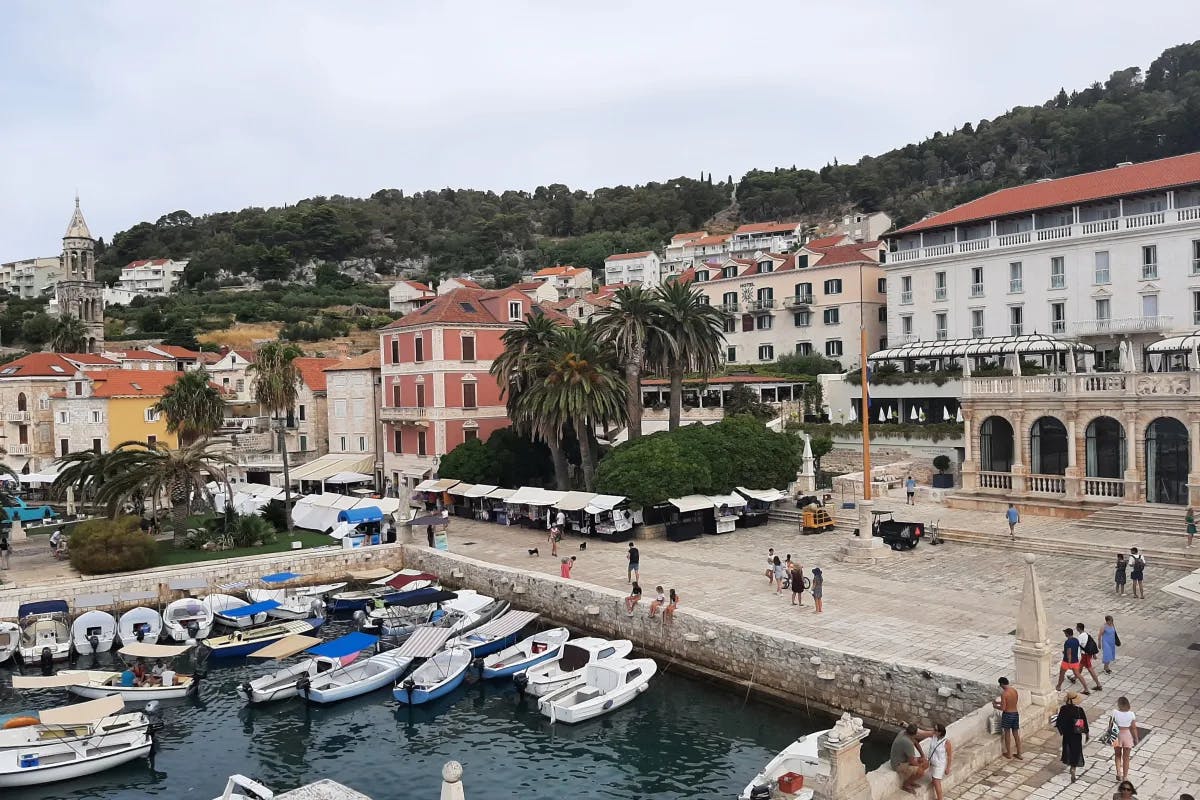 A photo of boats docked in a harbor near stone buildings with arches, red roofs and trees in the distance.