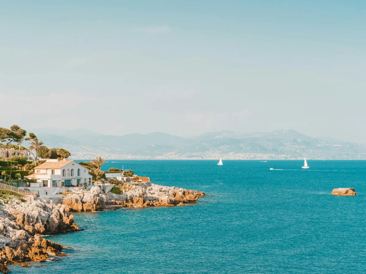 A tranquil coastal landscape with white buildings on a rocky shore, sailboats in the water, and distant hills under a clear sky.