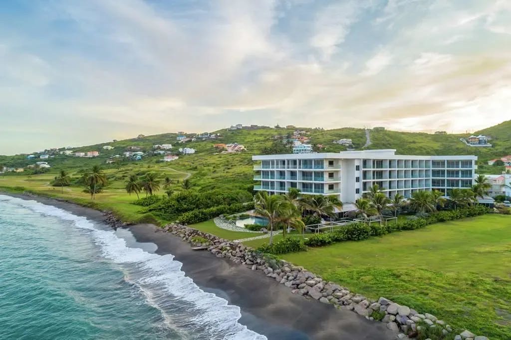 Turquoise waves crash on a volcanic beach separated from verdant fields that lead up to Koi Resort Saint Kitts, Curio Collection by Hilton