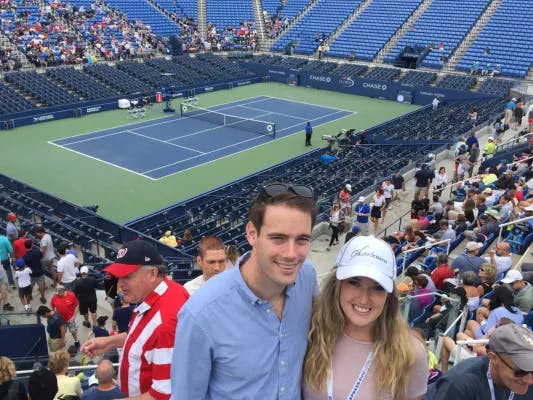 Hannah and a man posing in the stands with a tennis court behind them.