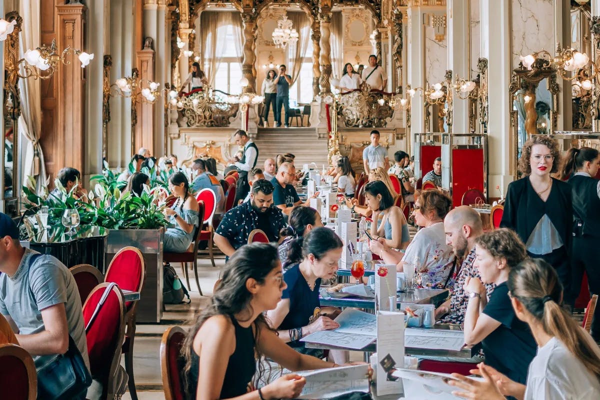 A luxurious café full of people dining around gold trimmed lights, red chairs, a staircase and potted plants.