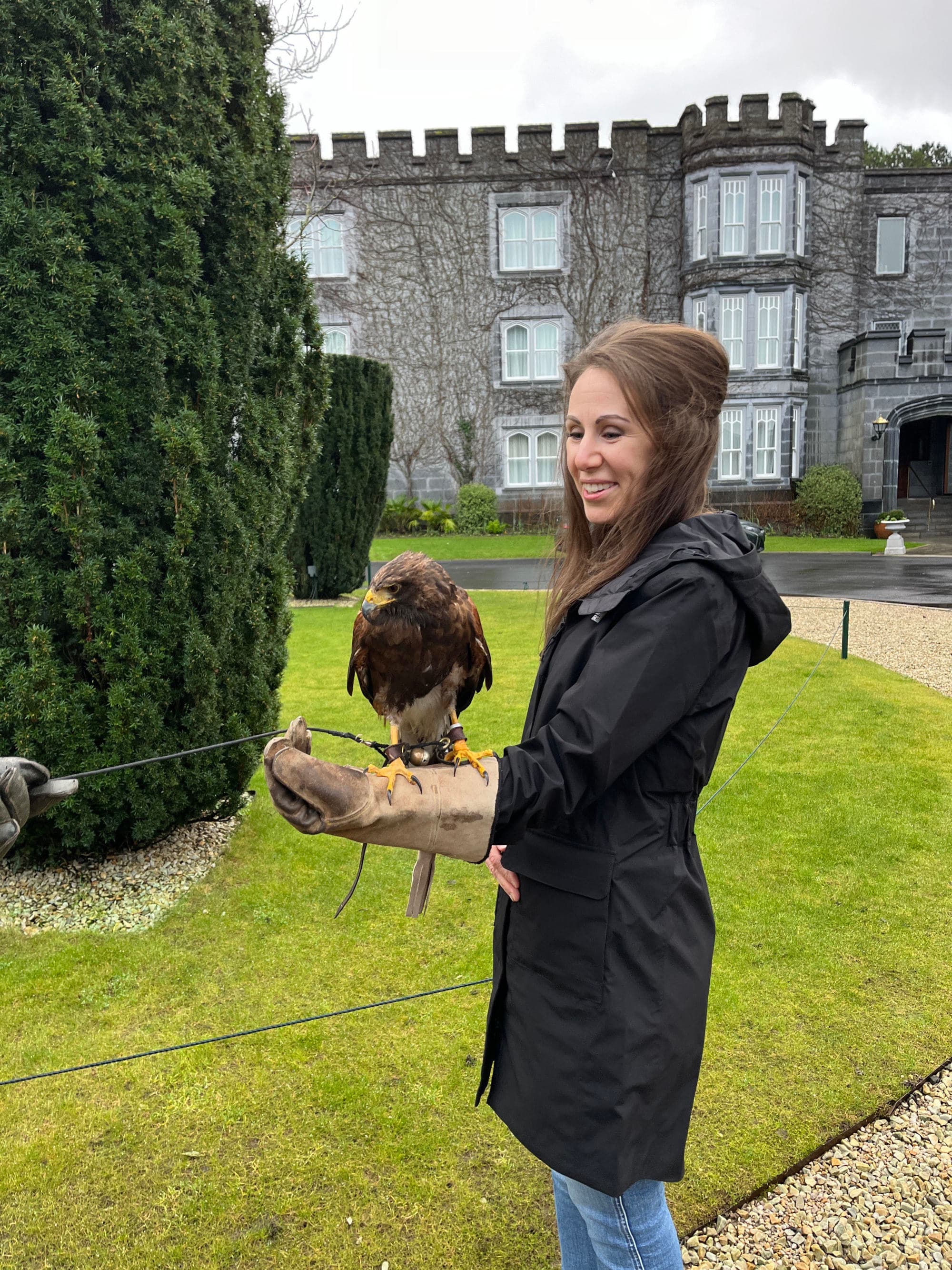 Megan standing in a jacket and blue jeans with a bird on their arm and castle in the background surrounded by green trees and grass. 