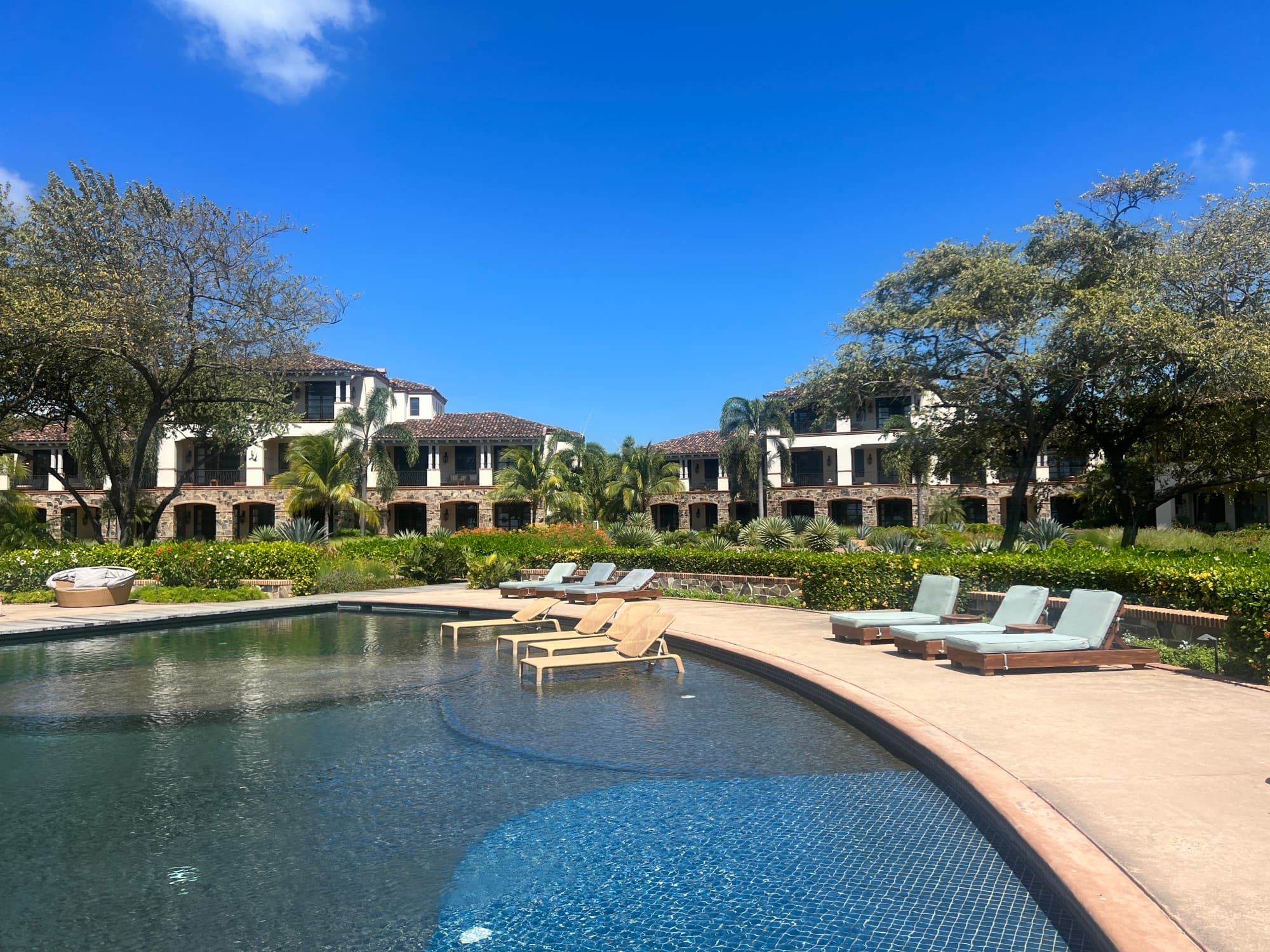 Pool area at a resort with sun loungers around the pool and some sun loungers in the pool, with green landscaping and two-storey buildings behind