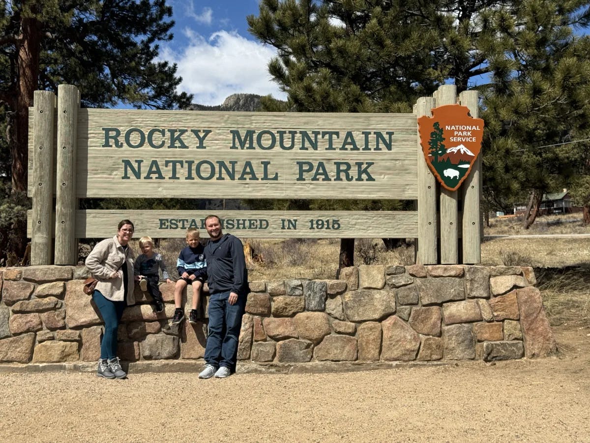 Travel advisor and family posing for a photo on a sunny day in front of the "rocky Mountain National Park" sign.