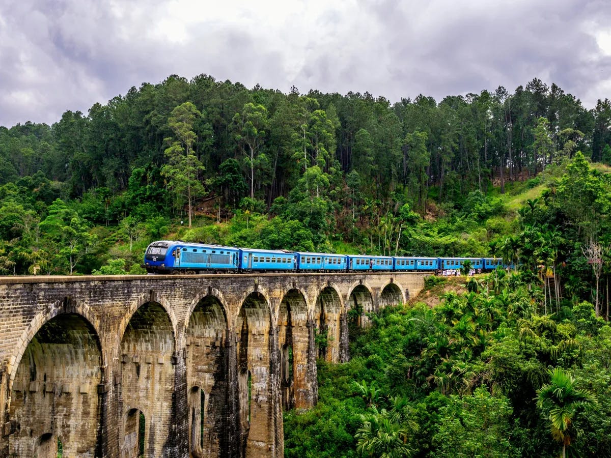 A blue train crosses an arched stone bridge surrounded by lush greenery under a cloudy sky.