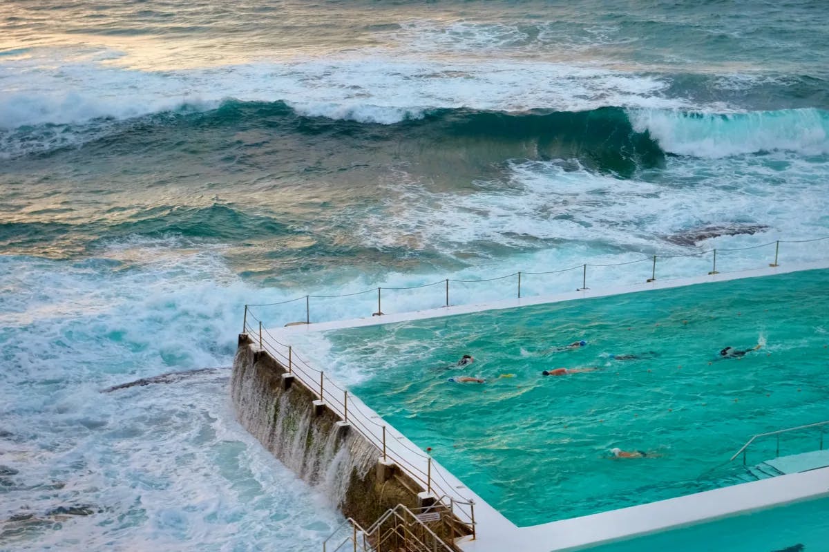 People swimming in a huge pool with the ocean waves crashing against it, located right on Bondi beach.