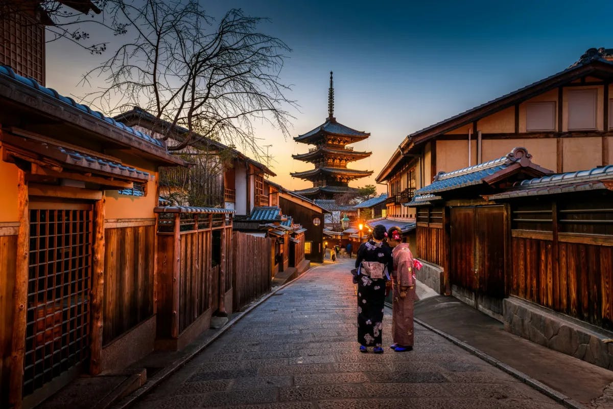 Two women in purple and pink kimonos standing on the street and looking out at a temple at sunset.