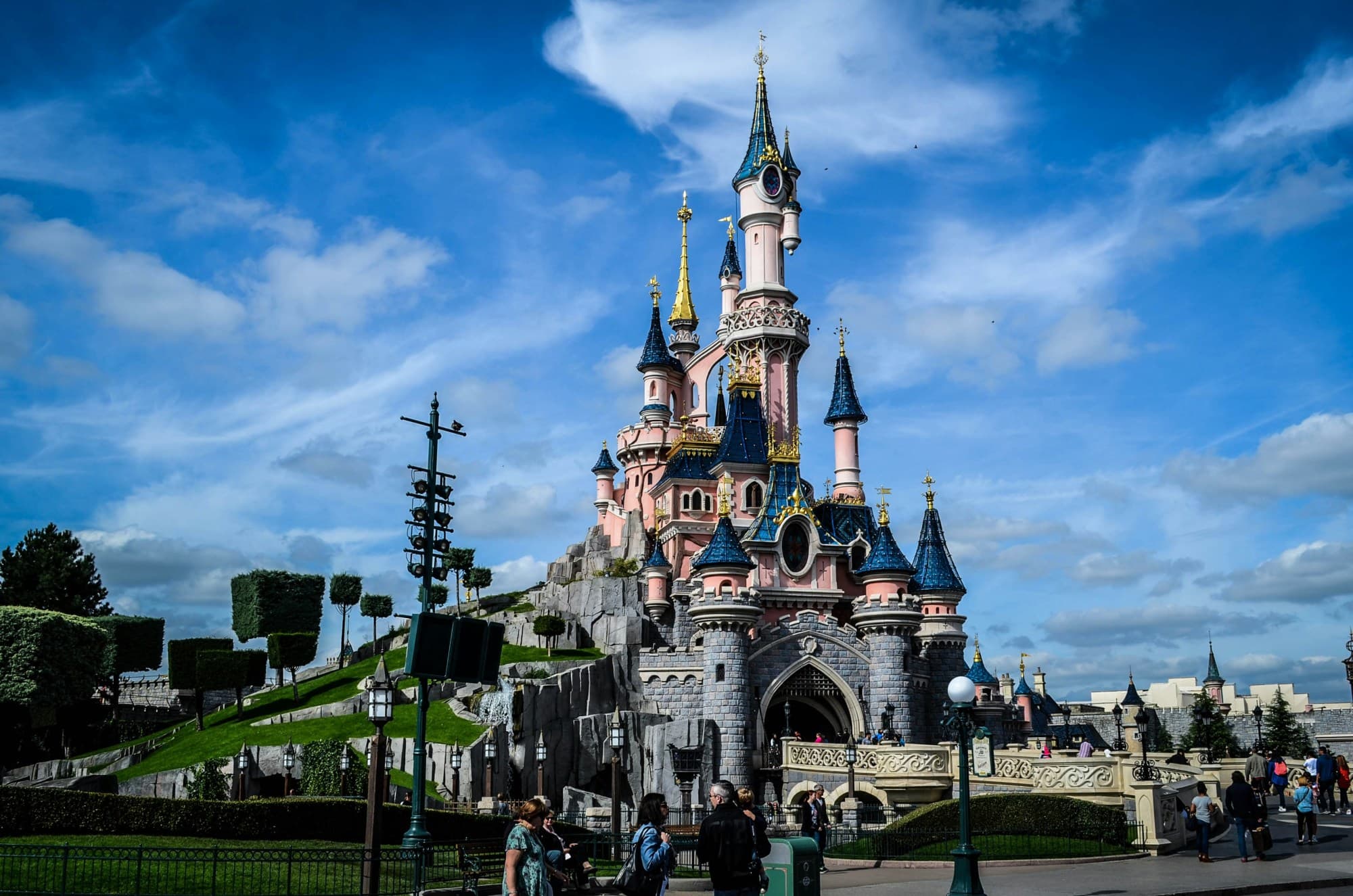 The Disney castle on a grassy hill with blue skies and clouds above it on a beautiful day.