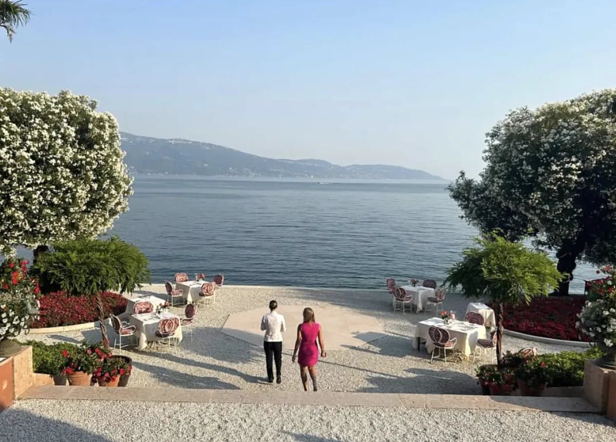 A view of a man and woman walking down a stone pathway to a beautiful lake with mountains in the distance. There are trees, potted flowers and dining tables in the surrounding areas. 