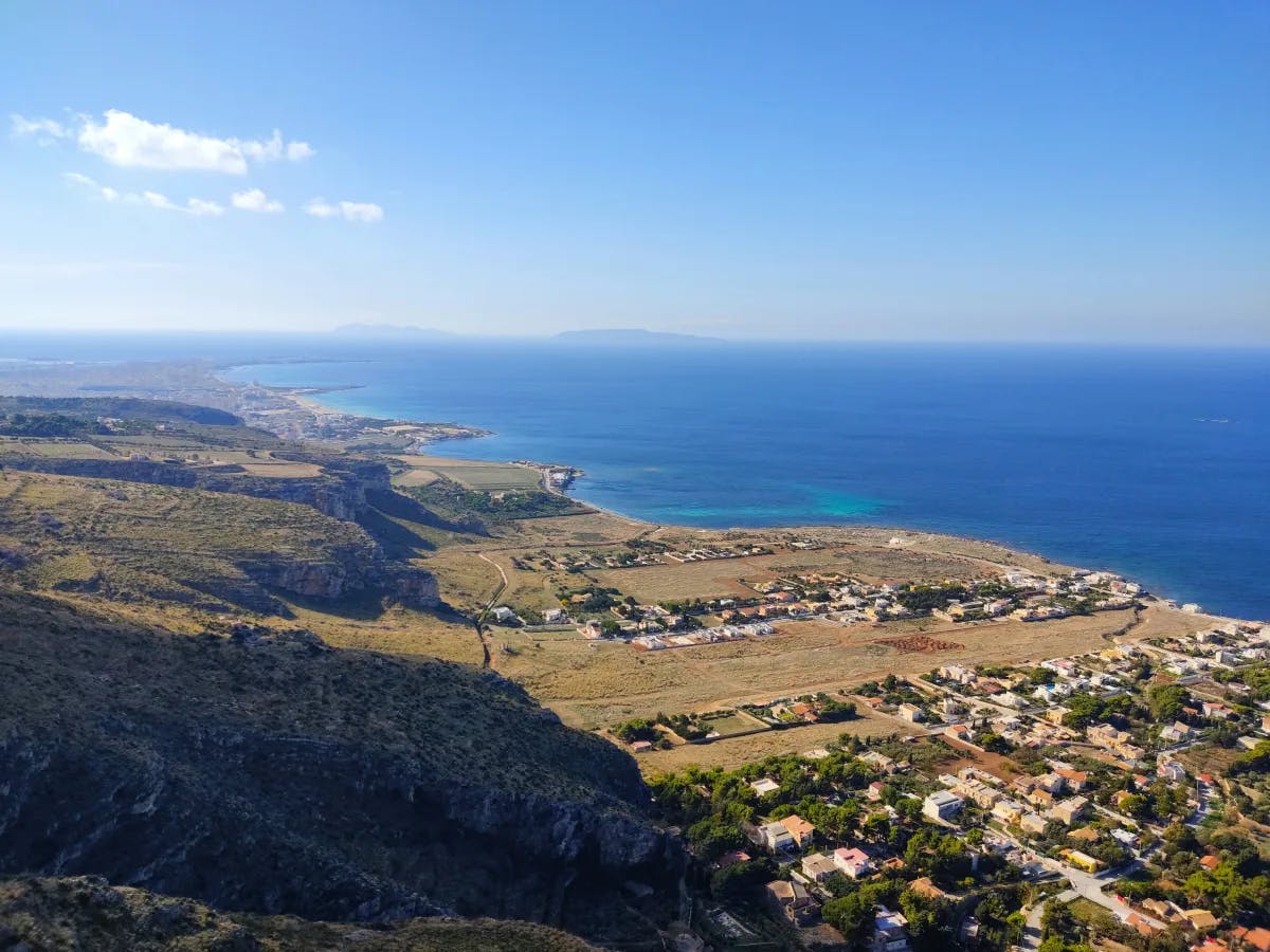 The image depicts a coastal landscape with a clear sky, overlooking a town by the sea with mountains in the background.