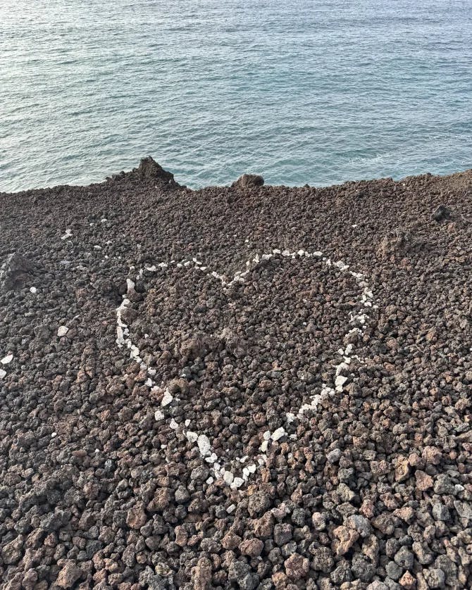 A heart made out of white pebbles on a rocky shore with blue water in the background.