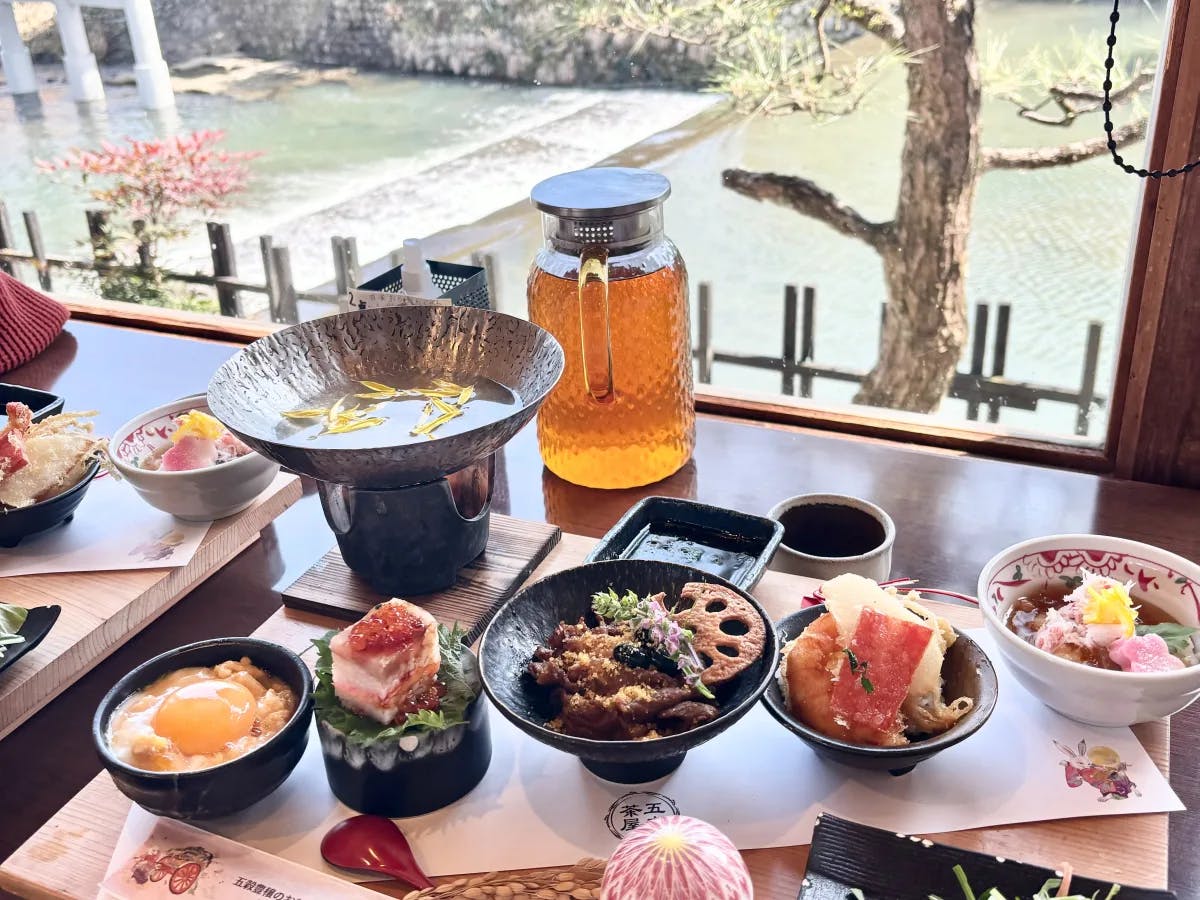 A picture of a table near a glass window overlooking a river, filled with traditional Japanese cuisine items.