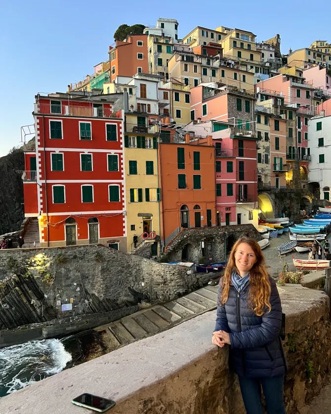 Kayla posing by a stone wall with colorful buildings visible behind her on a sunny day