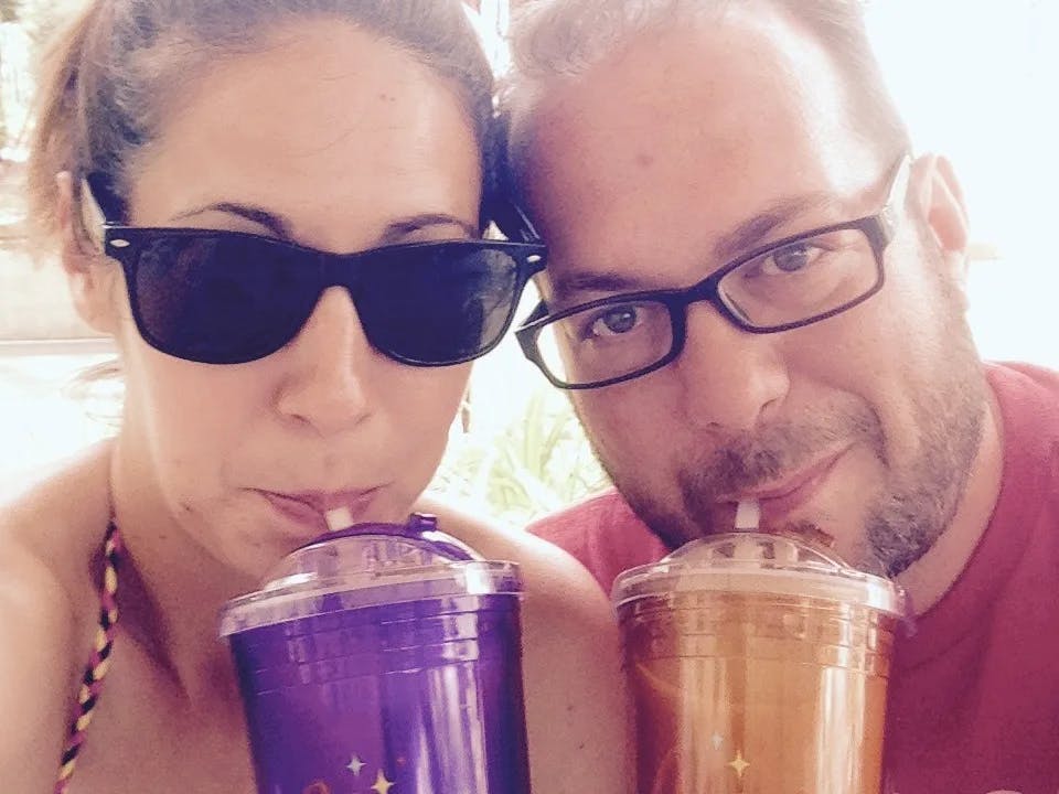 A man and woman posing for a selfie while sipping on cocktails outside. 