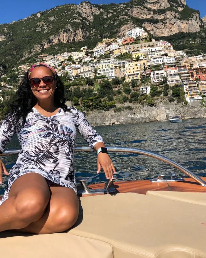 Shana sits on the front of a motor boat on the water of a coastal cliffside village