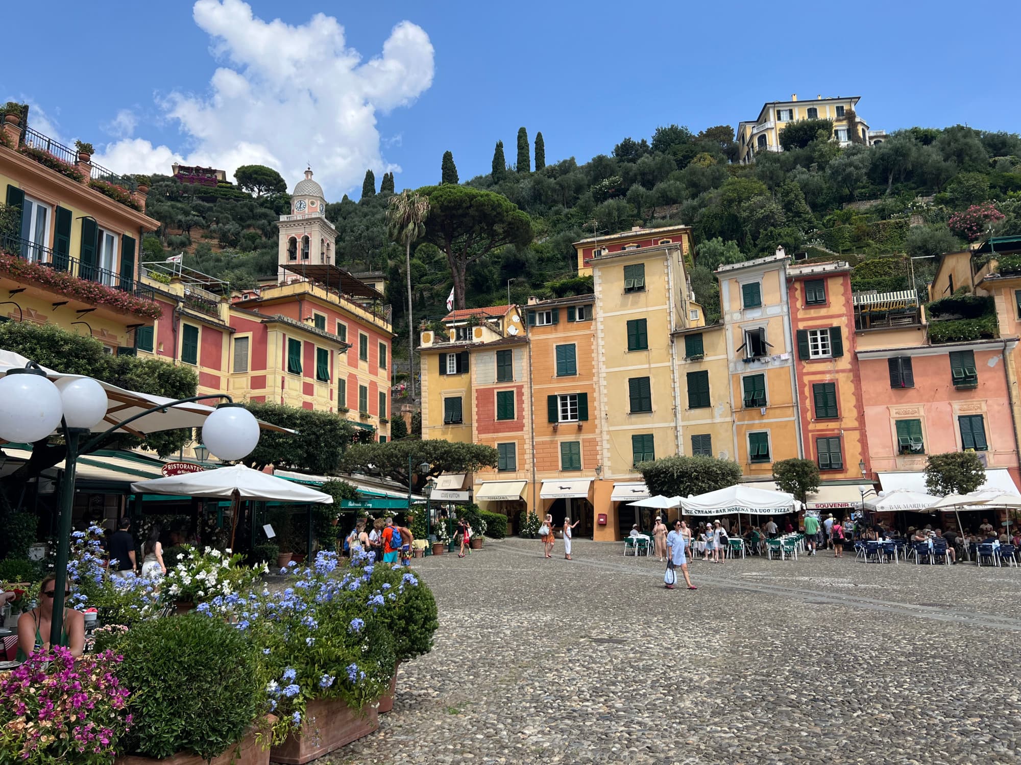 A view of a town center with people walking, lots of shops and tall, colorful buildings in a square at La Piazzetta outside the Splendido Mare DaV Mare restaurant.