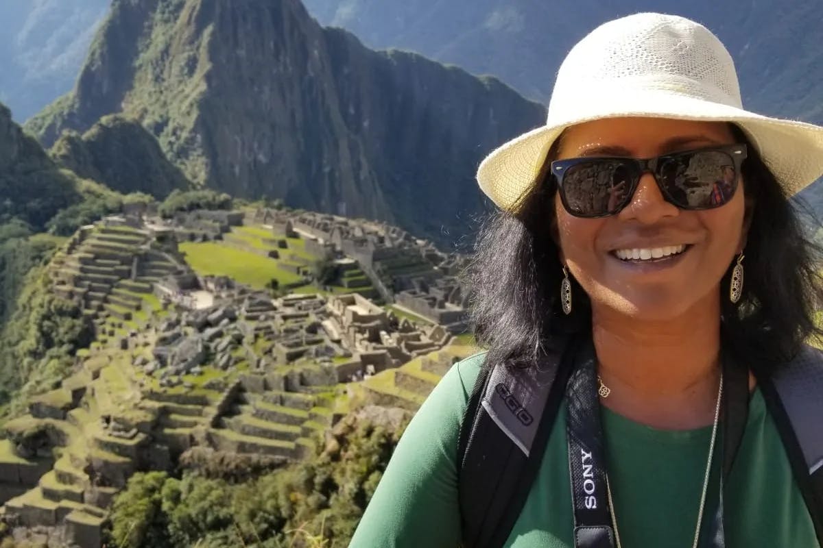 woman in green shirt and white hat standing with ruins and mountain in the background