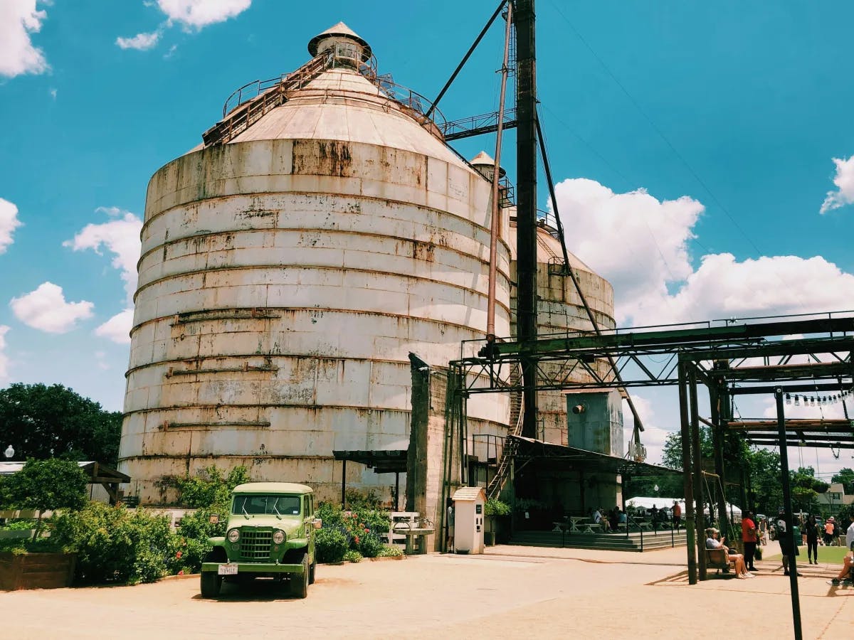 Two large industrial silos stand under a blue sky with scattered clouds, surrounded by metal structures and a vintage green truck in the foreground.