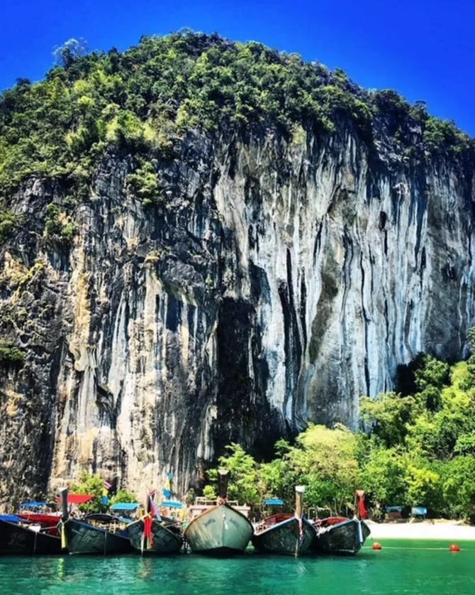 View of five fishing boats floating in front of a beach besides a large, lush green, steep cliff face