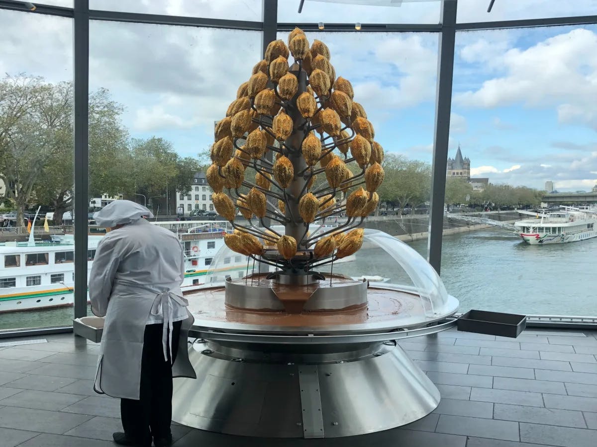 A large, golden pine cone sculpture is displayed inside a room with glass walls overlooking a body of water and a boat, with a person in white attire standing beside it.