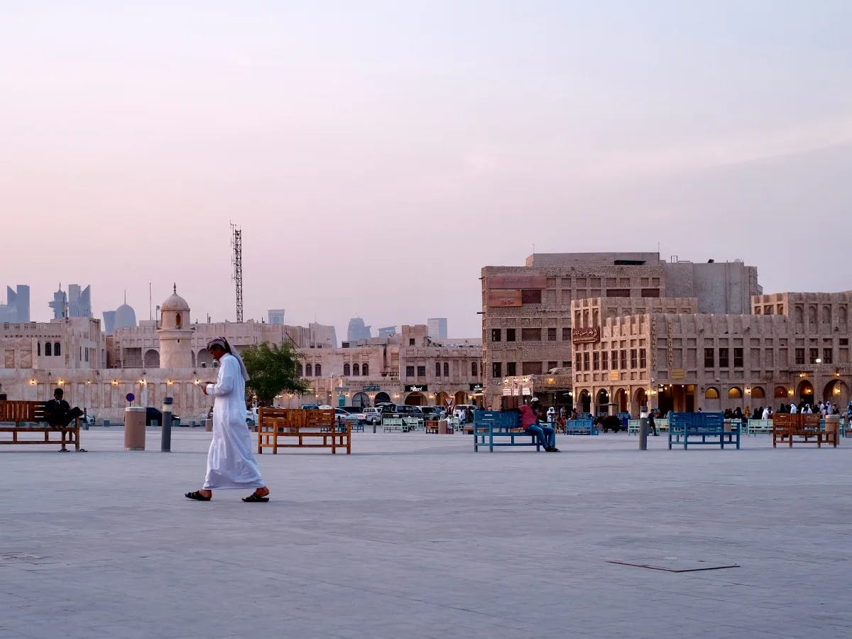 A person traverses a spacious square at dusk, flanked by traditional buildings and distant modern skyscrapers.