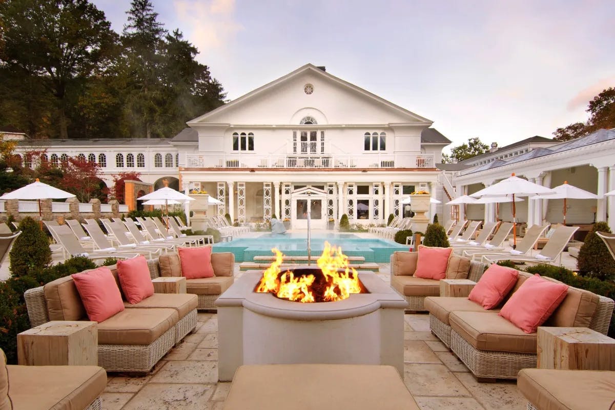 outdoor terrace with cushioned chairs and fire pit with pool and white exterior of hotel in the background